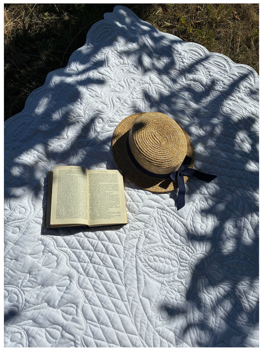 Couverture de lit blanche avec motif quilté, livre ouvert et chapeau en paille posé dessus, ombrages au soleil. Petite France, PETITE FRANCE. PHOTOGRAPHIE publicitaire.