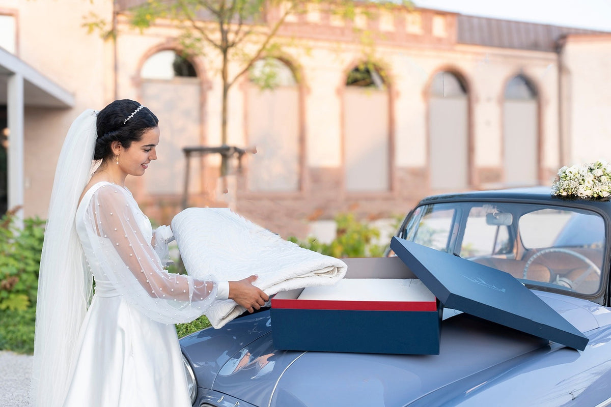Une mariée en robe blanche dépose une couverture dans une boîte cadeau sur le capot d’une voiture ancienne bleu gris, décor fleurs à l’arrière.