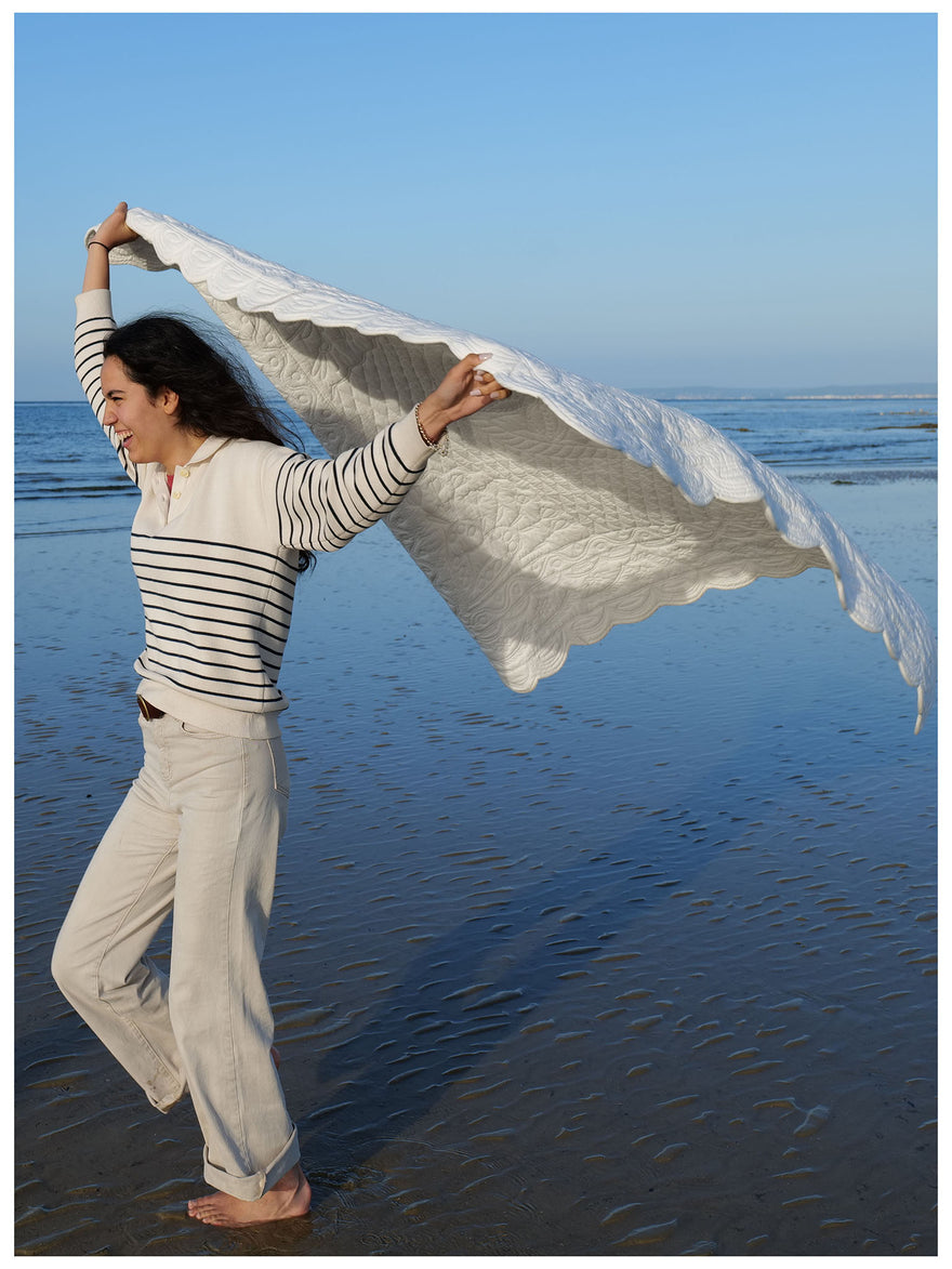 Homme souriant sur la plage porte un grand mouchoir ou voile blanc déployé comme cape, horizon mer bleu clair. Petite France.