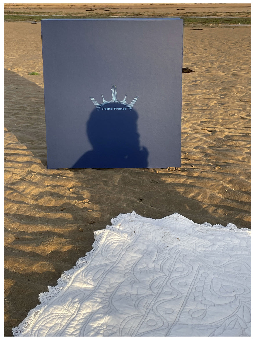 Une grande boîte bleue sur le sable affiche « Le carré de mariage Petite France » avec une ombre de personne devant. Un voile ou nappe blanche brodée est posé sur le sable au premier plan.