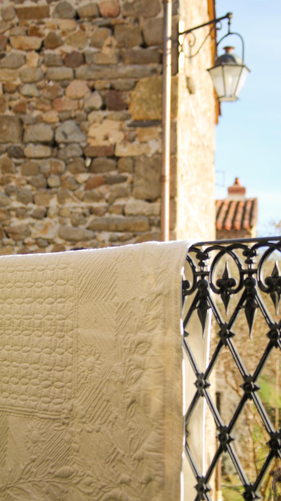 Dentelle blanche suspendue sur une balustrade en fer forgé devant un mur en pierre et une lanterne, style balcon provençal.