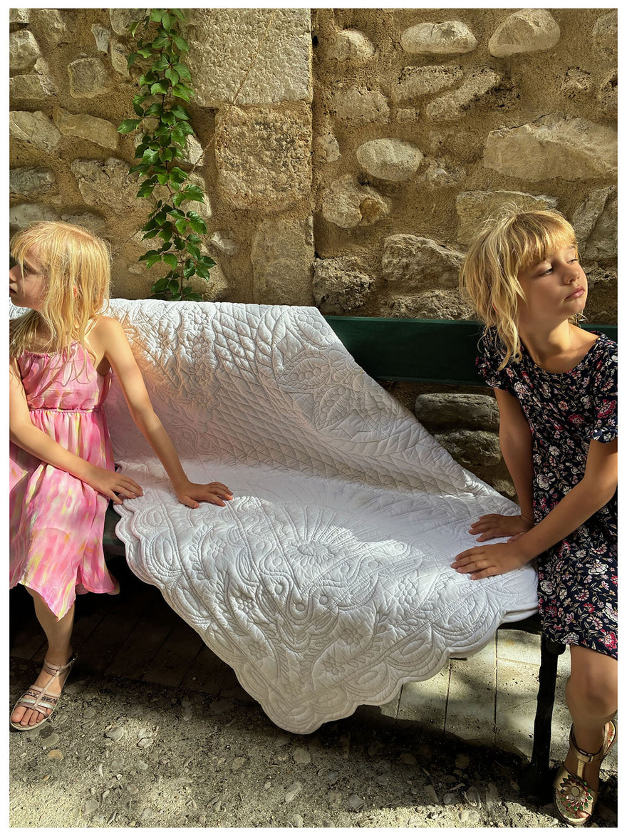 Deux jeunes filles assises sur un banc en pierre, tenant une grande couverture blanche texturée qui les couvre partiellement. Texte « Le car fé de mariage Petite France » en haut et « petite-france.fr » en bas.