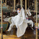 Jeune enfant sur cheval de manège blanc, drapée d’une grande étoffe blanche, au milieu d’une grande roue. Décor coloré et structure dorée de manège, ambiance festive. 
