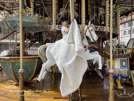 Jeune enfant sur cheval de manège blanc, drapée d’une grande étoffe blanche, au milieu d’une grande roue. Décor coloré et structure dorée de manège, ambiance festive. "The French wedding throw by Petite France" visible.
