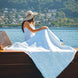 Femme assise sur une barque en bois sur l’eau, chapeau large, manteau blanc et serviette blanche couvrant l’avant, paysage lacustre et collines au loin.