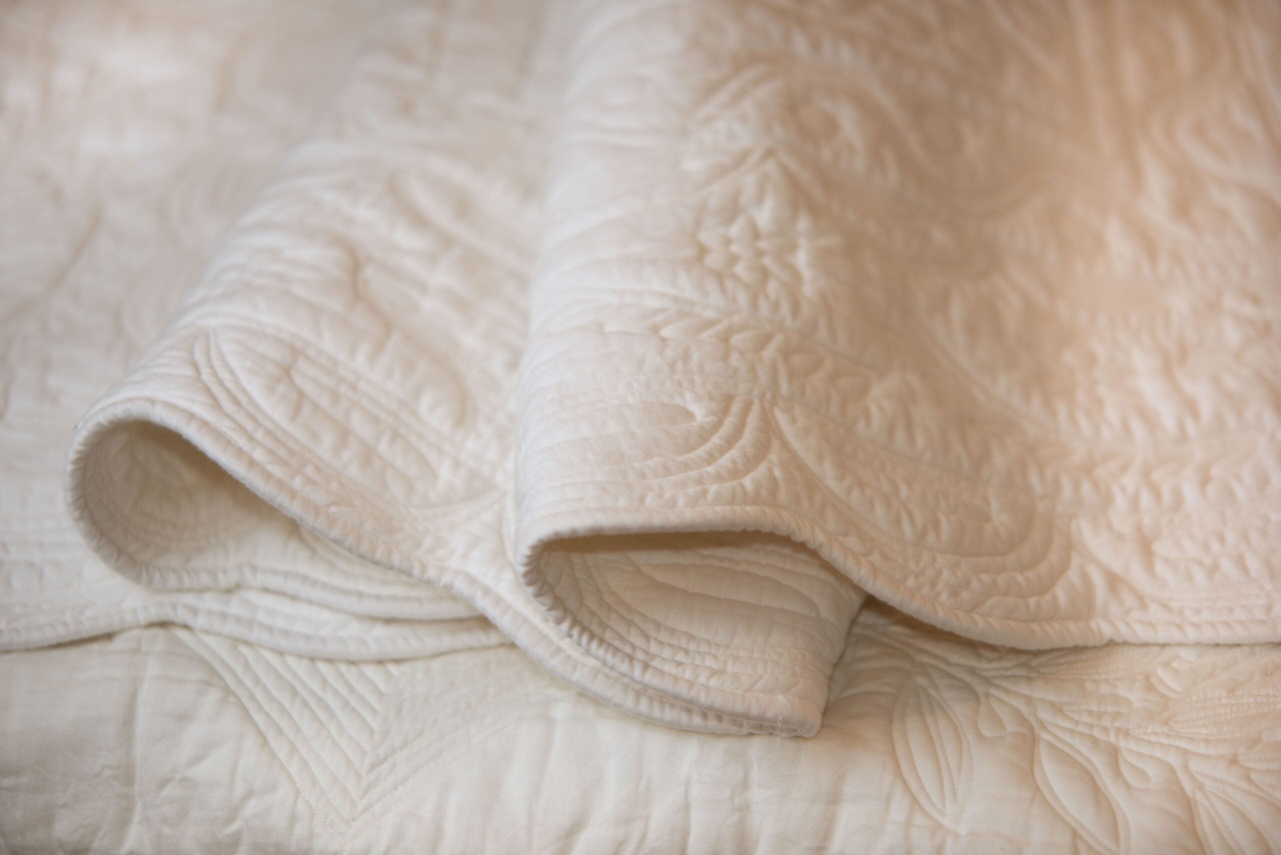 Jeté de lit beige avec motif quilté texturé et bord roulé, posé sur une surface plane.