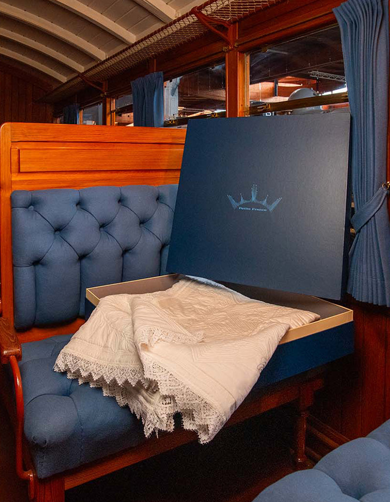 Coffre ouvert sur un coussin bleu et un drap blanc à dentelle, posé sur un canapé-tiroir bleu dans un wagon bois.