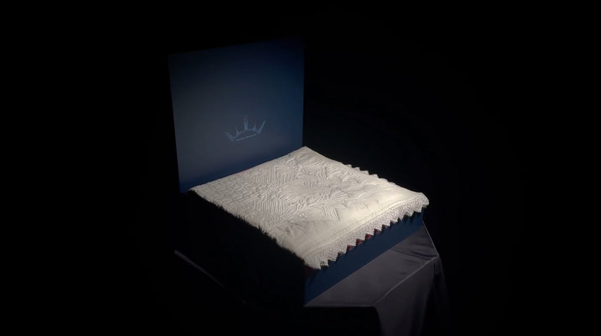 Couverture blanche brodée sur un lit ou socle bleu foncé, éclairée au centre dans l’obscurité.
