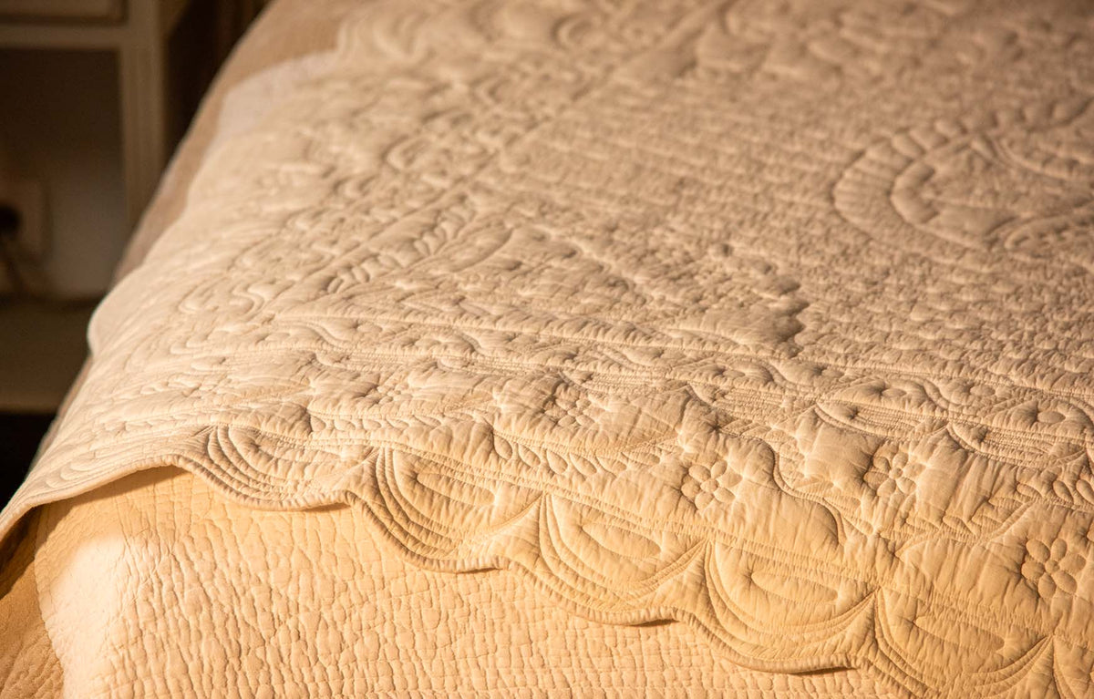 Lit enteint par la lumière chaude avec couverture beige à motifs dentelle, texture quiltée et surpiqûres visibles.