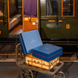 Deux valises et deux boîtes bleues sur une charrette métallique devant un wagon de train, côté quai, lumière chaude.