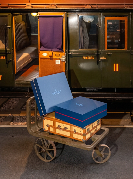 Deux valises et deux boîtes bleues sur une charrette métallique devant un wagon de train, côté quai, lumière chaude.