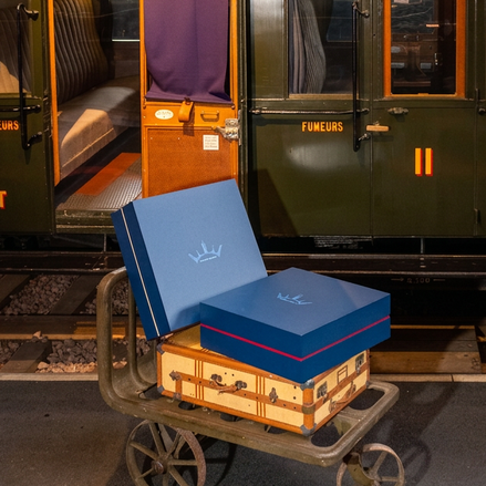Deux valises et deux boîtes bleues sur une charrette métallique devant un wagon de train, côté quai, lumière chaude.