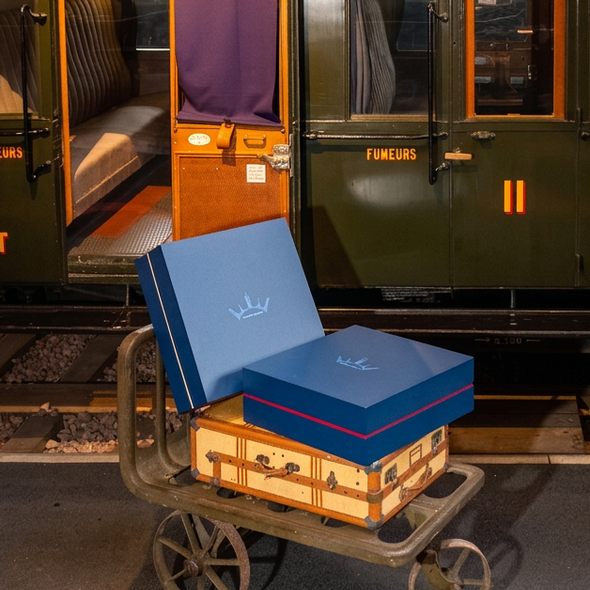 Deux valises et deux boîtes bleues sur une charrette métallique devant un wagon de train, côté quai, lumière chaude.