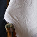 Couverture blanche ornée de motifs floraux et dentelle posée sur un fauteuil à bois et tissu décoratif.