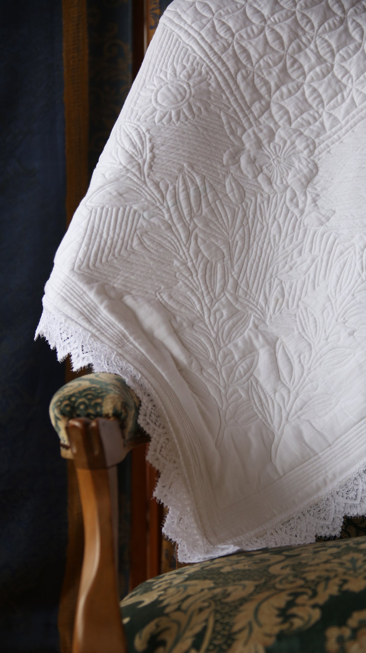 Couverture blanche ornée de motifs floraux et dentelle posée sur un fauteuil à bois et tissu décoratif.