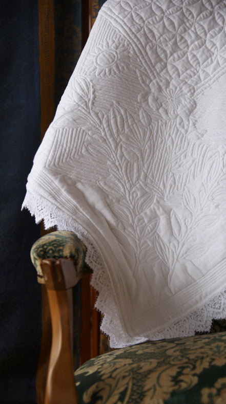 Couverture blanche ornée de motifs floraux et dentelle posée sur un fauteuil à bois et tissu décoratif.