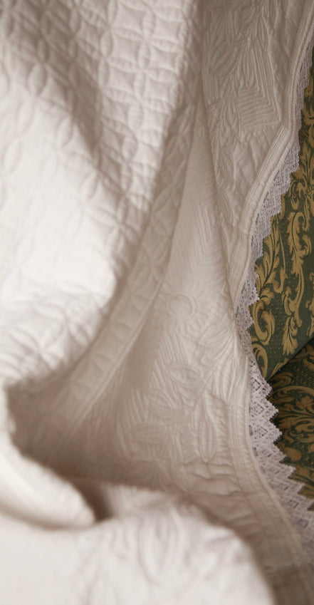 Voilage ou couverture blanche à motif discret, bordure en dentelle près d’un tissu d’ameublement vert et doré.