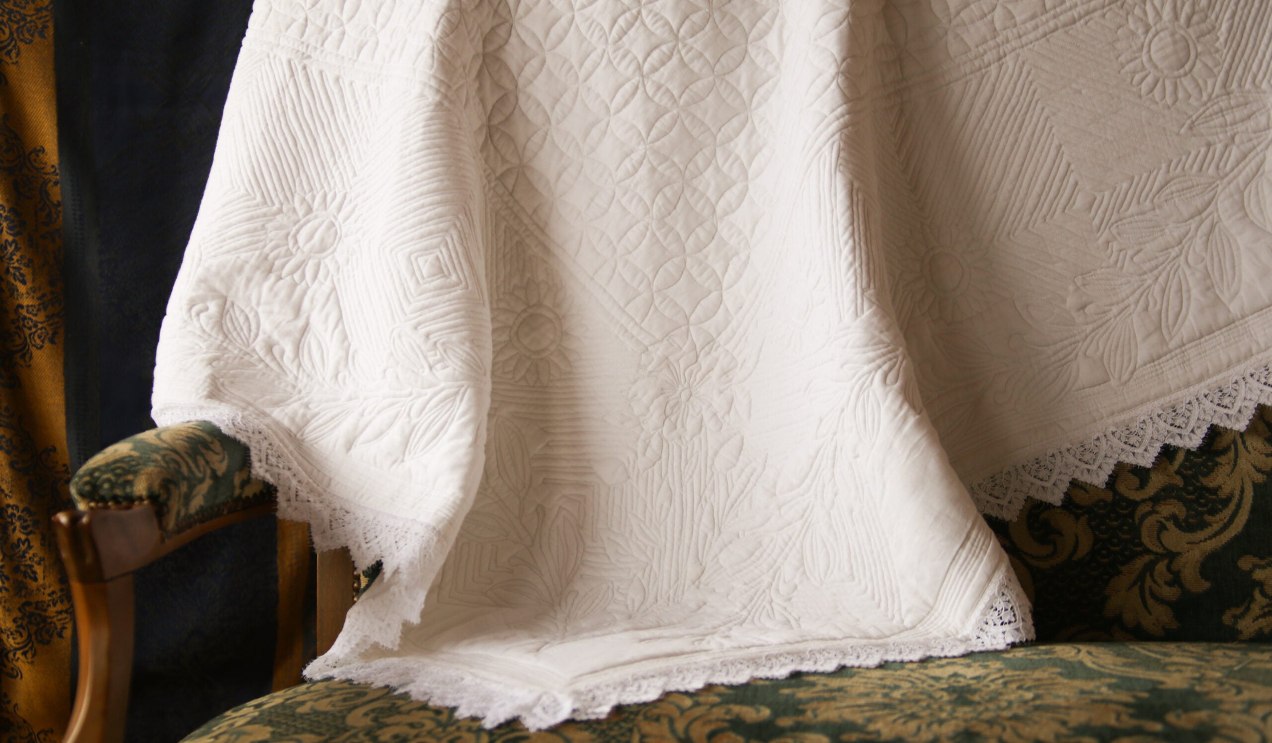 Couverture en dentelle blanche légère posée sur un canapé vert et doré à motif, avec bordures festonnées visibles.