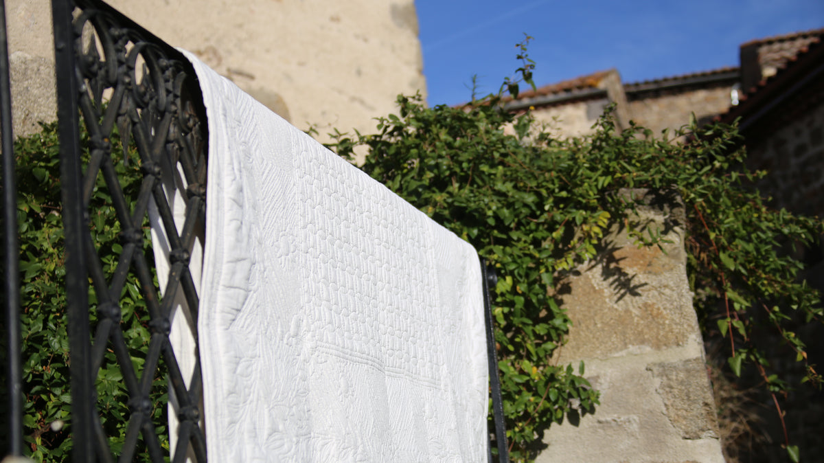 Linge blanc accroché au-dessus d’une barrière métallique, séchant au soleil sur fond de mur et de végétation.