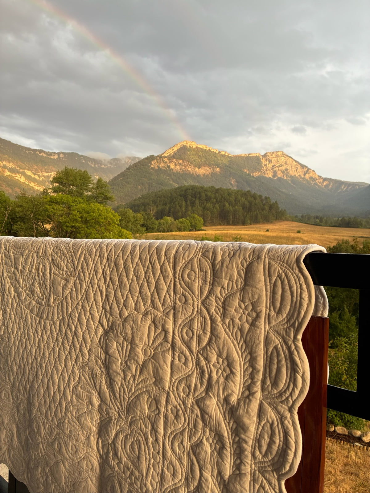 Couverture quiltée blanche drapée sur une rambarde avec vue sur montagnes, prairie et arc-en-ciel tôt le matin.