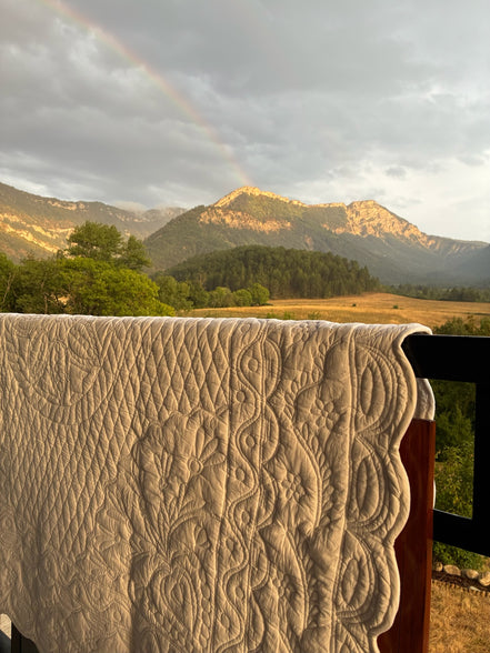 Couverture quiltée blanche drapée sur une rambarde avec vue sur montagnes, prairie et arc-en-ciel tôt le matin.