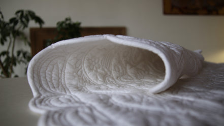 Drap housse blanc matelassé plié, textures quiltées visibles et conforment sur une table.