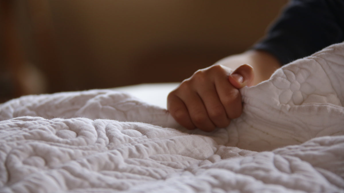 Mains d’un enfant sur une couverture quiltée, doigts serrés près du bord du drap blanc.