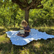 Une jeune fille lit un livre assise sur une grande couverture blanche posée sur l’herbe sous un arbre en plein air.