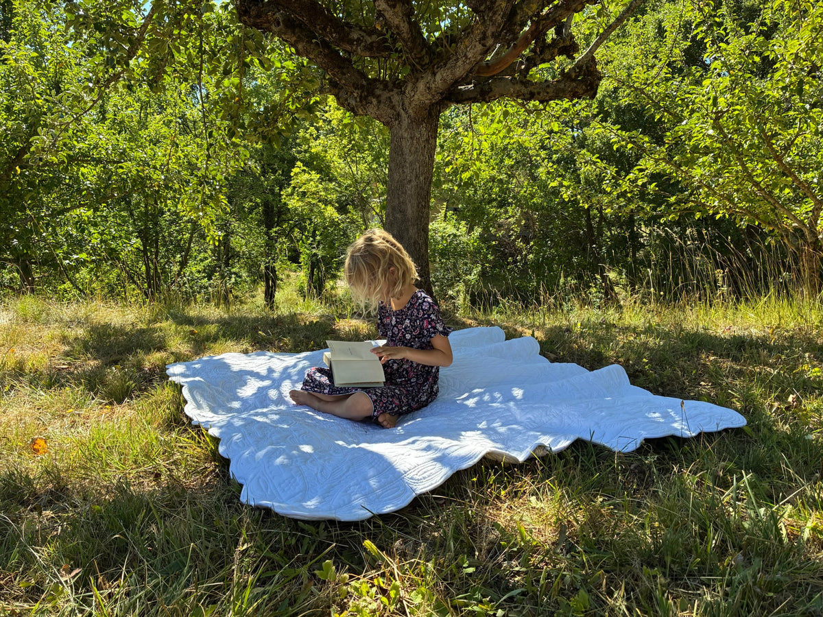 Une jeune fille lit un livre assise sur une grande couverture blanche posée sur l’herbe sous un arbre en plein air.