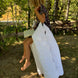 Fille assise en plein air, pieds nus, portant un foulard floral et une grande couverture blanche qu’elle utilise comme cape. Elle tient un petit livre ouvert sur les genoux sous les arbres.