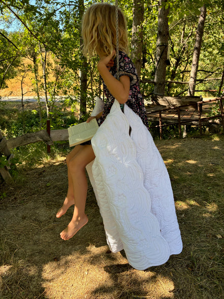 Fille assise en plein air, pieds nus, portant un foulard floral et une grande couverture blanche qu’elle utilise comme cape. Elle tient un petit livre ouvert sur les genoux sous les arbres.