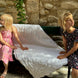 Deux jeunes filles assises sur un banc en pierre près d’un mur en pierres, tenant une grande couverture blanche ornée, l’une en robe rose et l’autre en robe à motifs, sous une lumière estivale.
