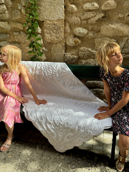 Deux jeunes filles assises sur un banc en pierre près d’un mur en pierres, tenant une grande couverture blanche ornée, l’une en robe rose et l’autre en robe à motifs, sous une lumière estivale.