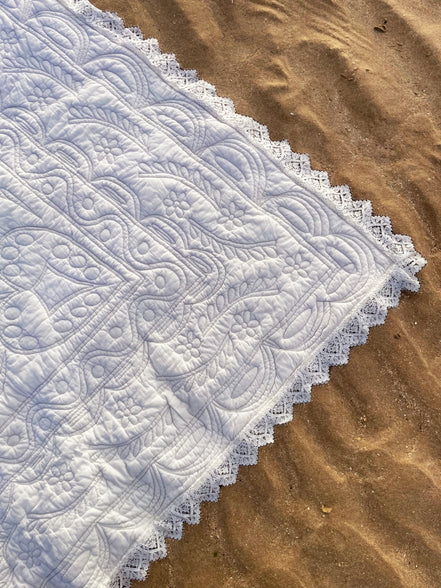 Dentelle blanche à motifs floraux et géométriques posée sur sable, bordure en dentelle fine.