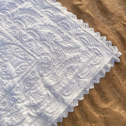 Dentelle blanche à motifs floraux et géométriques posée sur sable, bordure en dentelle fine.