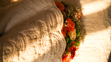 Arrière-plan floral au milieu d’une couette blanche à motifs, avec bouquet de roses orange et autres fleurs blanches et rouges éclairé par une lumière chaude.