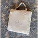 Sac fourre-tout beige avec poignées, posé sur un tapis à motifs gris. Logo bleu discret en son centre.