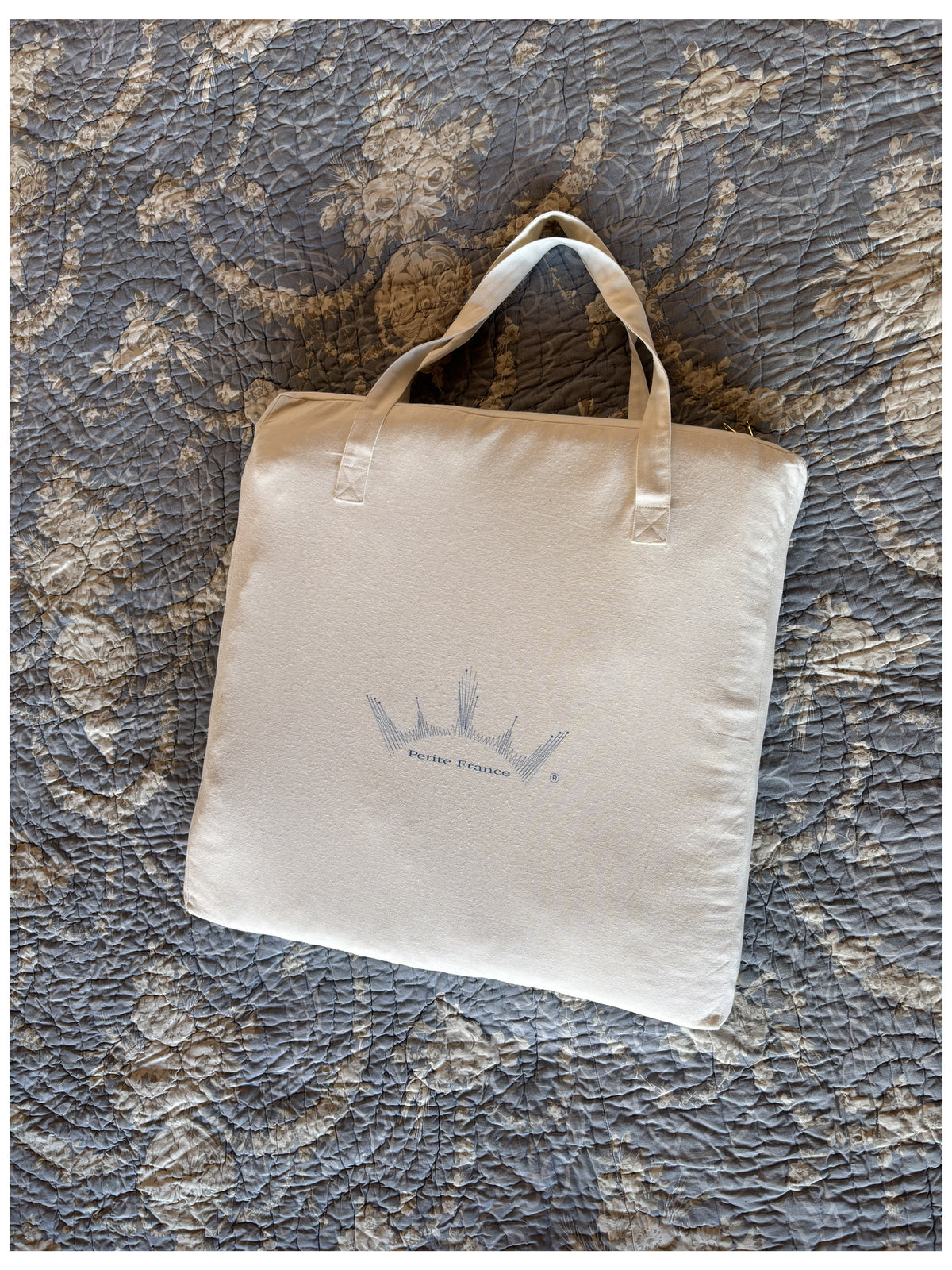 Sac fourre-tout beige avec poignées, posé sur un tapis à motifs gris. Logo bleu discret en son centre.