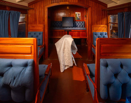 Intérieur d'un wagon-restaurants ou compartimentées en bois, banquettes bleues, tissu tufté et rideaux bleus, avec une couverture blanche posée sur le sol.