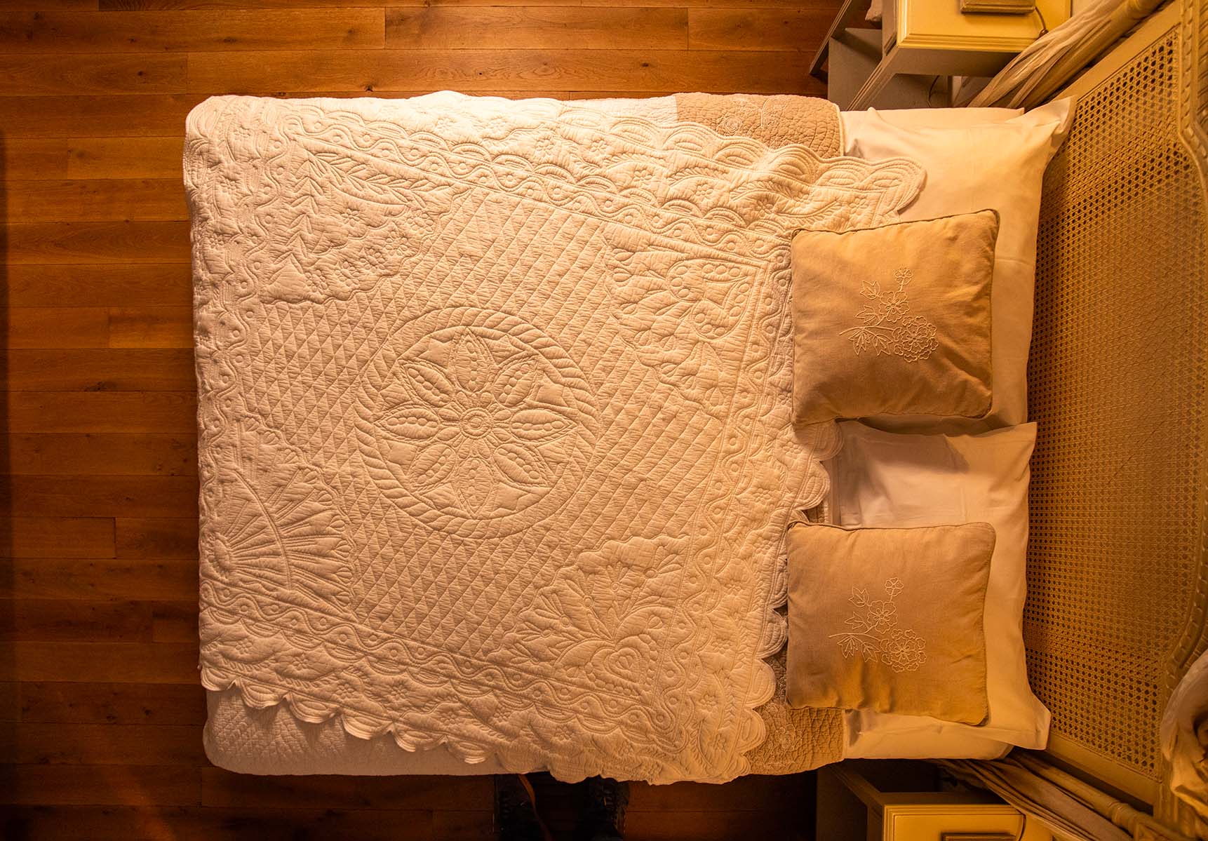 Lit double vu du dessus, avec drap blanc et couverture ornée, deux gros oreillers beige et une tête de lit en osier.