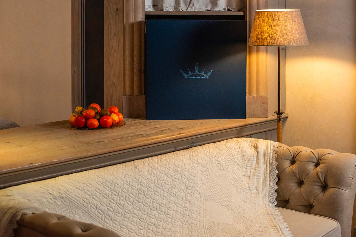 Livré dans un intérieur cosy: un grand livre bleu sombre posé sur une tablette, près d’une lampe, et un petit plat de tomates cerises sur le bois, canapé beige et couverture blanche en tissage.