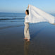Femme debout sur la plage, tenant une grande serviette blanche déployée derrière elle, au bord de l’eau calme. Le ciel est clair et l’océan s’étend à l’horizon.