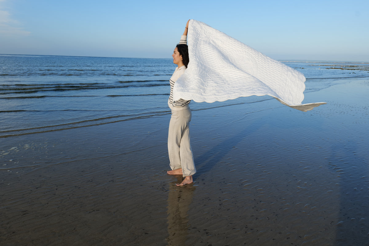 Femme debout sur la plage, tenant une grande serviette blanche déployée derrière elle, au bord de l’eau calme. Le ciel est clair et l’océan s’étend à l’horizon.