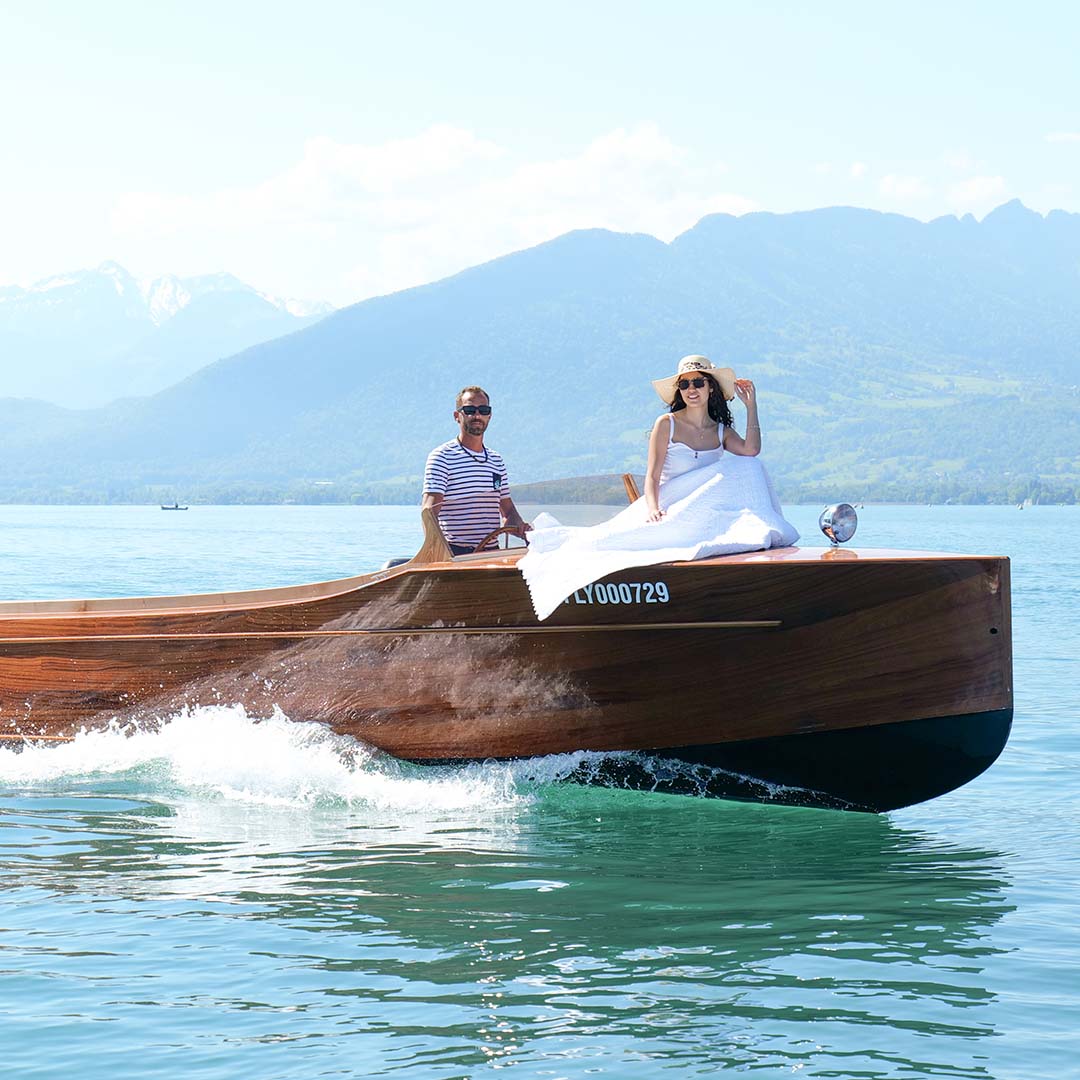 Deux personnes en bateau en bois sur un lac calme, avec femmes en robe blanche et chapeau, montagne en fond.