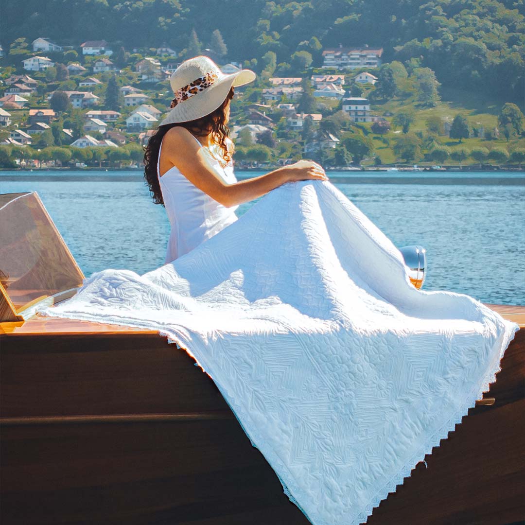 Femme en robe blanche et chapeau, assise sur un bateau au bord de l’eau, couvrant le pont avec une grande couverture blanche. Vue sur une rive avec maisons verdoyantes.