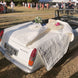 Voiture décapotable blanche décorée de linge et fleurs pour un mariage en plein air, avec tentes et invités en arrière-plan.
