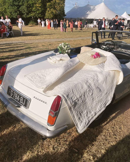 Voiture décapotable blanche décorée de linge et fleurs pour un mariage en plein air, avec tentes et invités en arrière-plan.