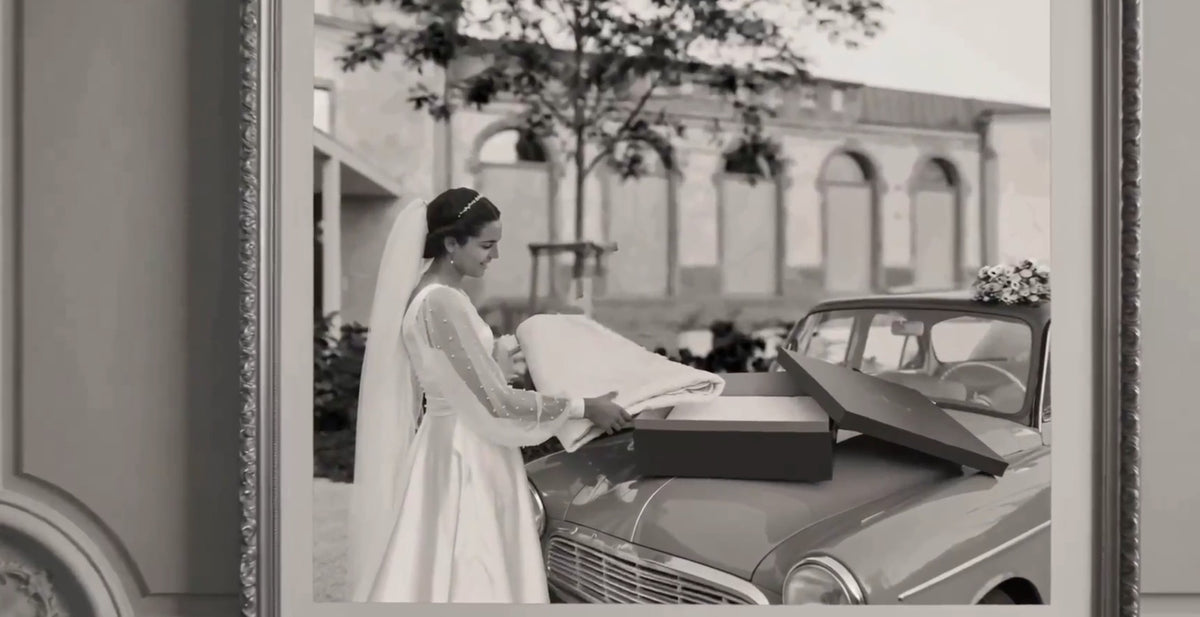 Une jeune mariée en robe blanche dépose un bouquet ou un objet sur une voiture décapotable grise lors d’un mariage, près d’un cadre décoratif.