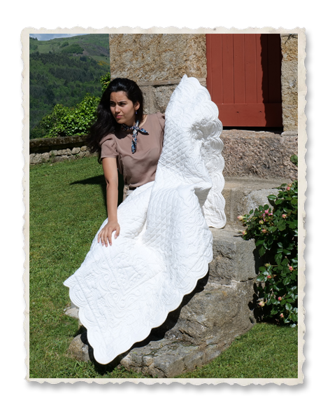 Femme assise sur une bordure en pierre, portant une grande nappe blanche décorative ou couverture quiltée devant un bâtiment en pierre. Jardin et paysage vert en arrière-plan.
