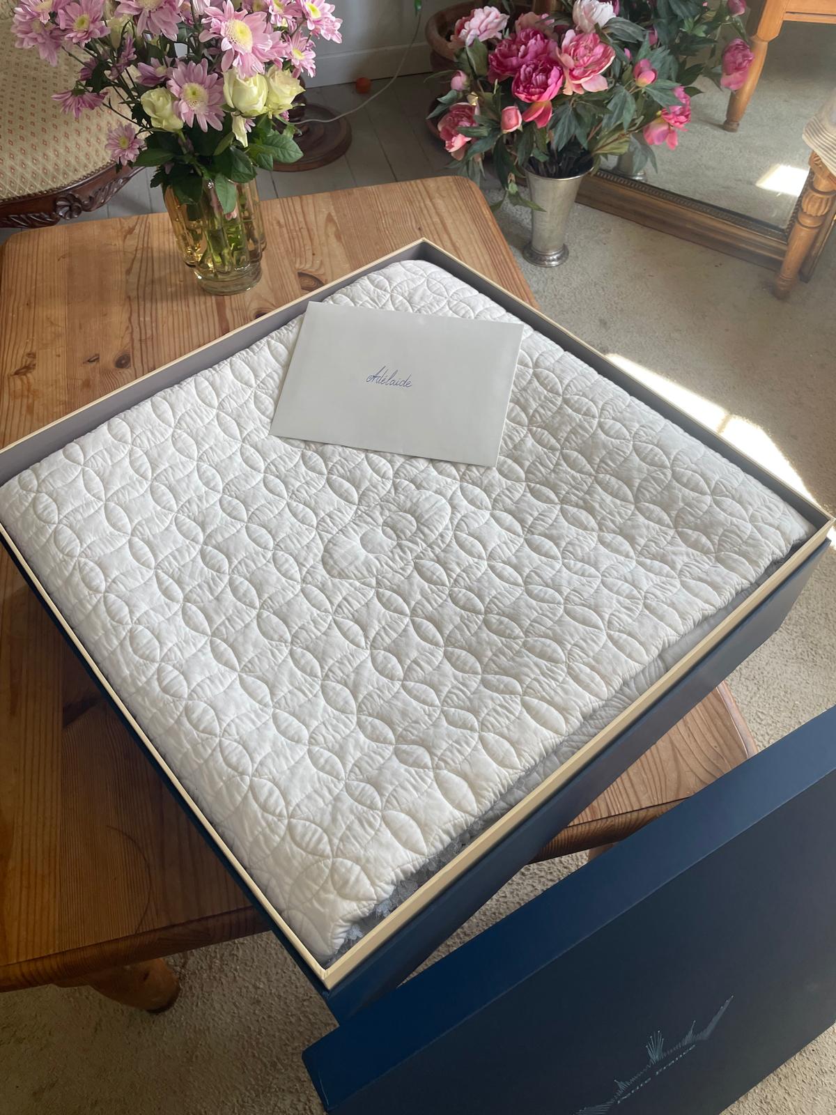 Matelas matelassé blanc dans une boîte cadeau bleu, posé sur une table en bois, avec des bouquets de fleurs au fond.